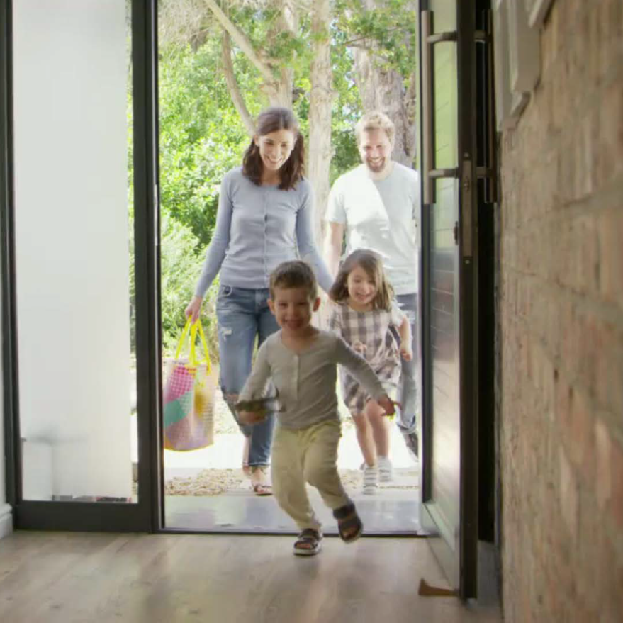 A family walk into a house smiling, two children running, carrying groceries